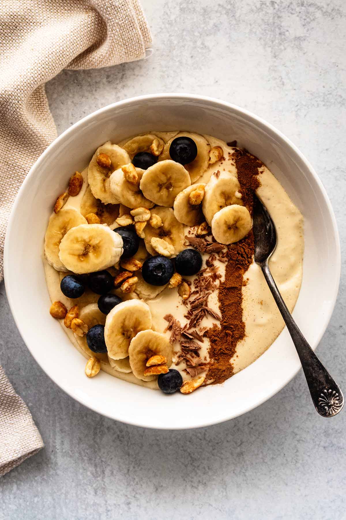 Banana smoothie bowl topped with chopped peanuts, fresh blueberries, chocolate shavings, and a line of ground cinnamon in a white bowl with a spoon.