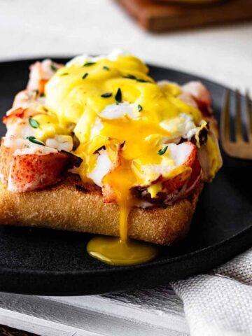 Lobster benedict with yolk dripping down on a dark grey plate with a gold fork.