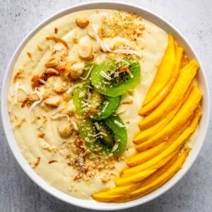 Mango smoothie bowl in a white bowl topped with chopped macadamia nuts, sliced kiwi, fresh mango slices, and toasted coconut flakes on a concrete countertop.