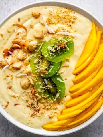 Mango smoothie bowl in a white bowl topped with chopped macadamia nuts, sliced kiwi, fresh mango slices, and toasted coconut flakes on a concrete countertop.