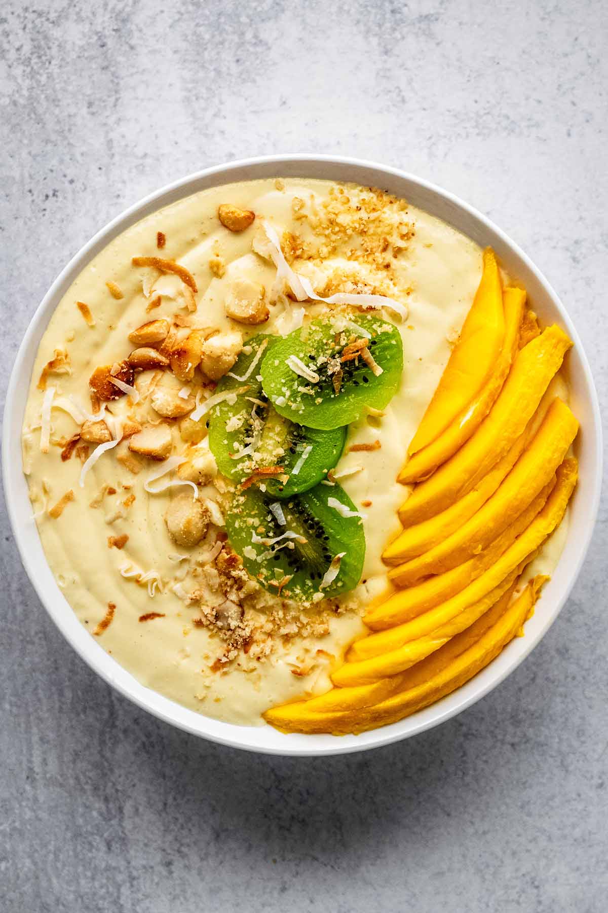 Mango smoothie bowl in a white bowl topped with chopped macadamia nuts, sliced kiwi, fresh mango slices, and toasted coconut flakes on a concrete countertop.