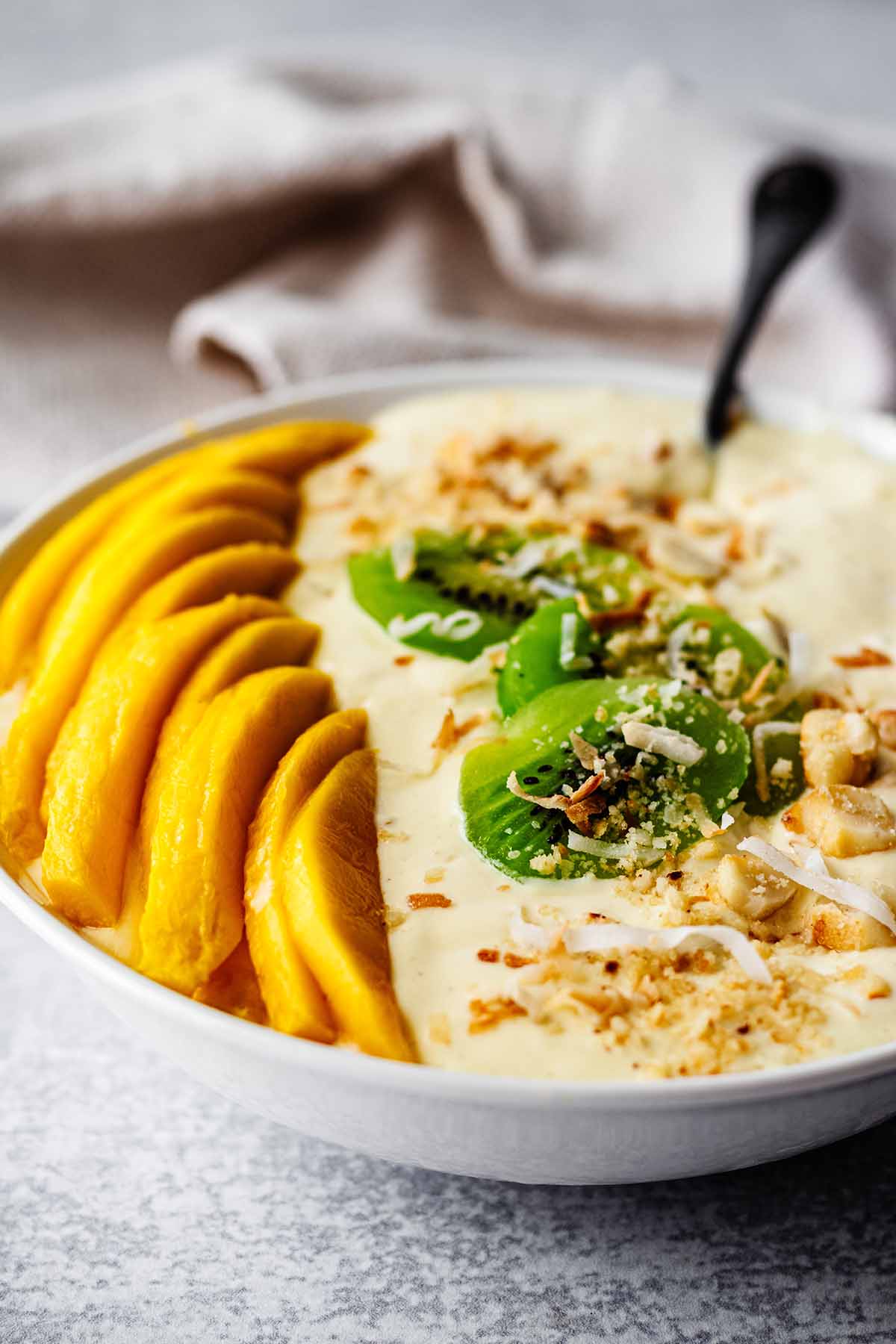 Front view of thick mango smoothie in a white bowl topped with fresh mango slices, chopped macadamia nuts, sliced kiwi, and toasted coconut flakes with a spoon and a light beige napkin.