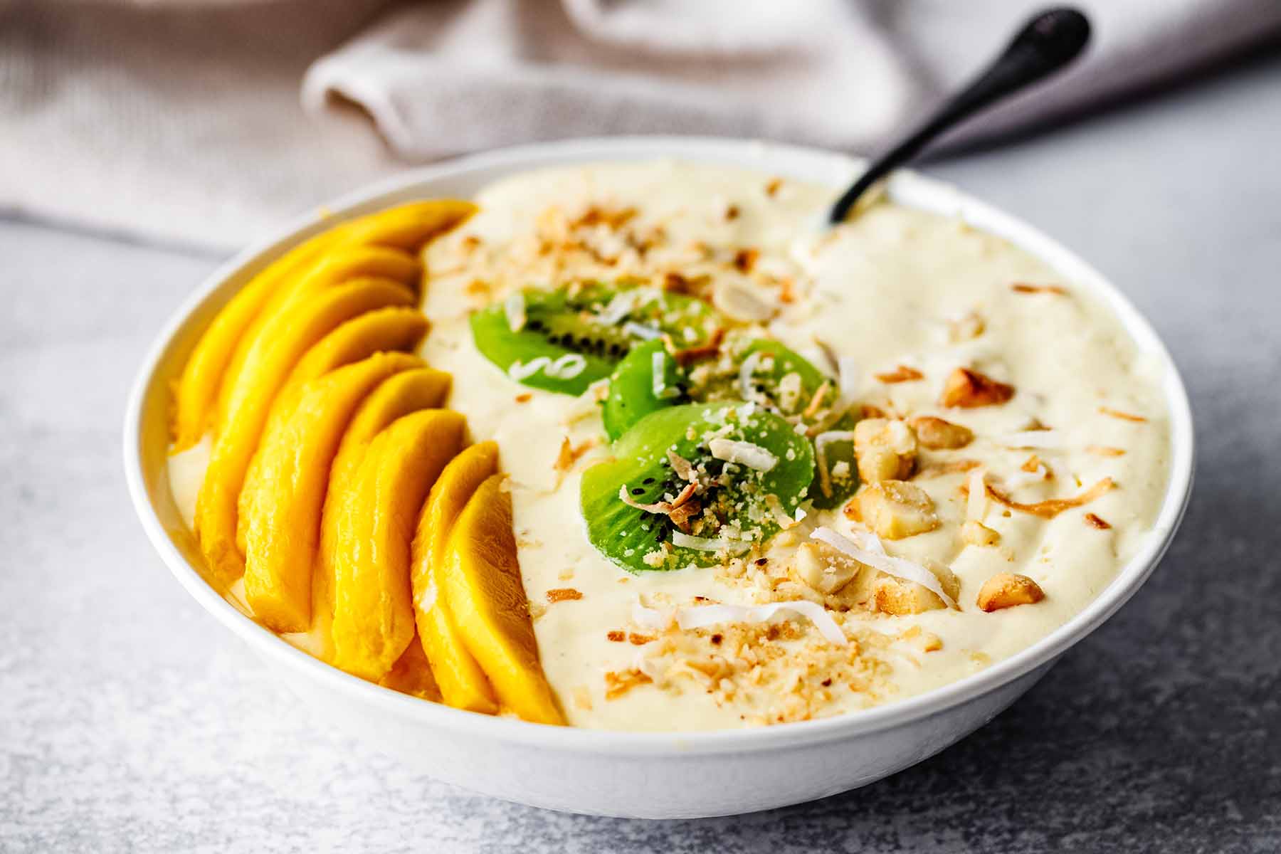 Mango smoothie bowl topped with sliced kiwi and mango, chopped toasted macadamia nuts, and toasted coconut flakes in a white bowl with a spoon. 