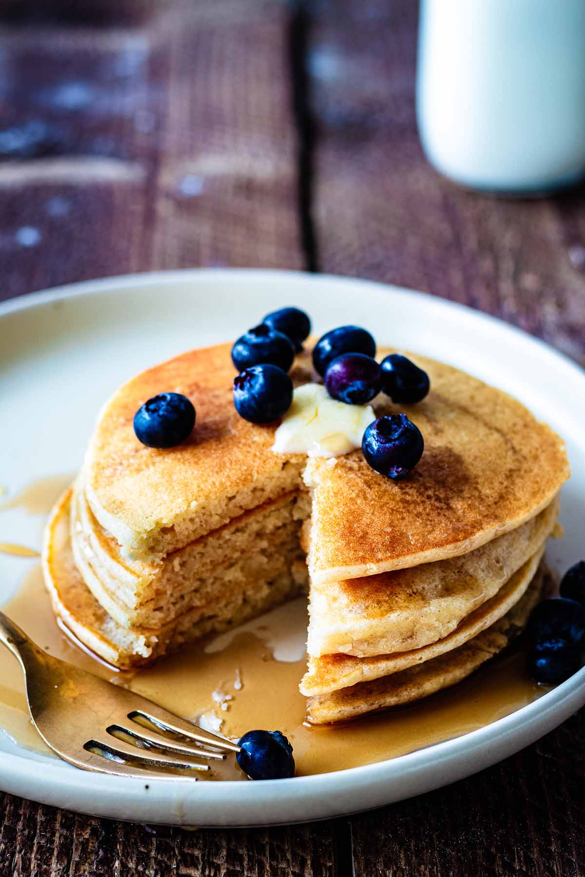 Short stack of cassava pancakes topped with butter, fresh blueberries, and maple syrup on a beige plate with a gold fork.