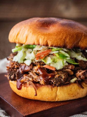Crockpot shredded beef sandwich on a dark wood cutting board on top of a cream napkin.