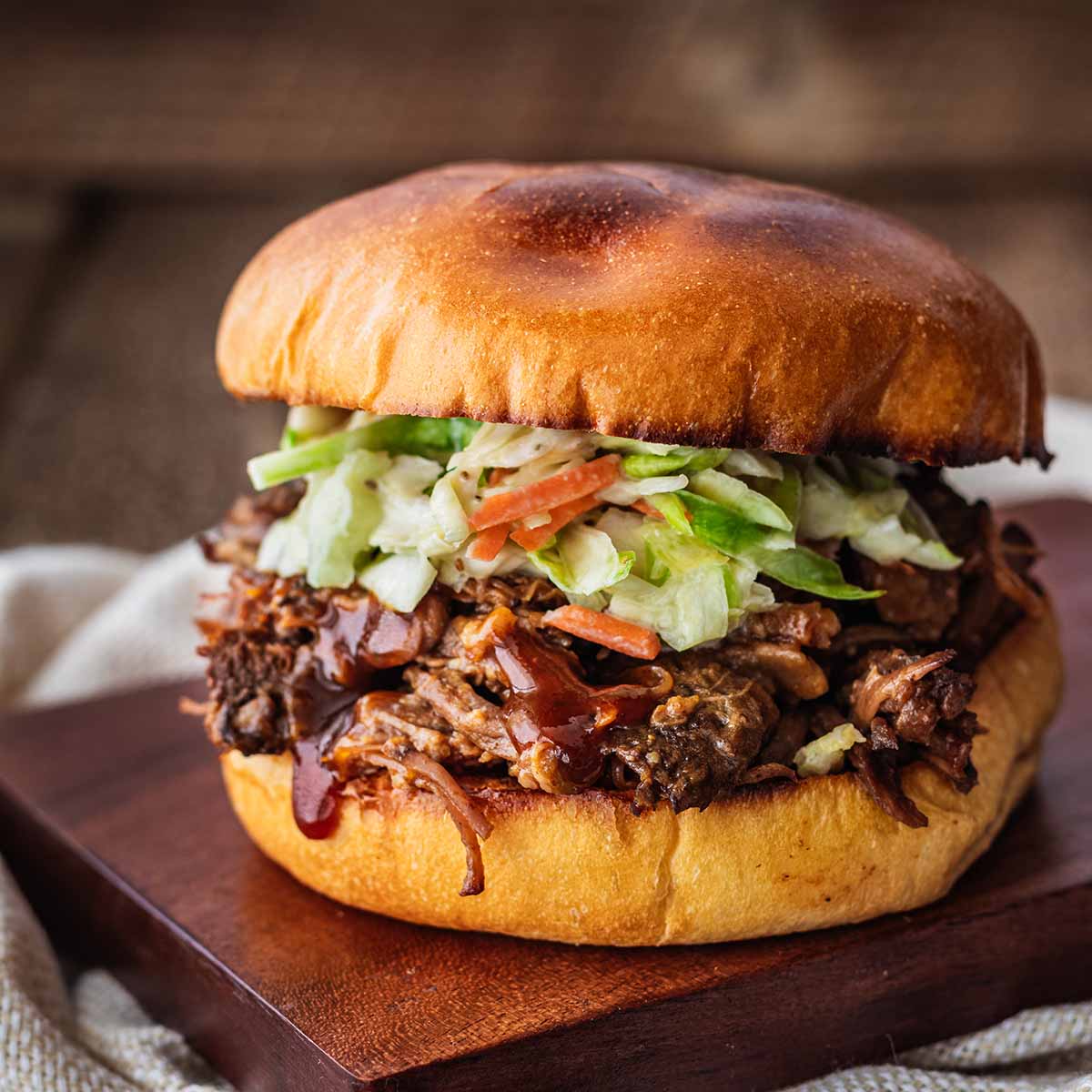 Crockpot shredded beef sandwich on a dark wood cutting board on top of a cream napkin.