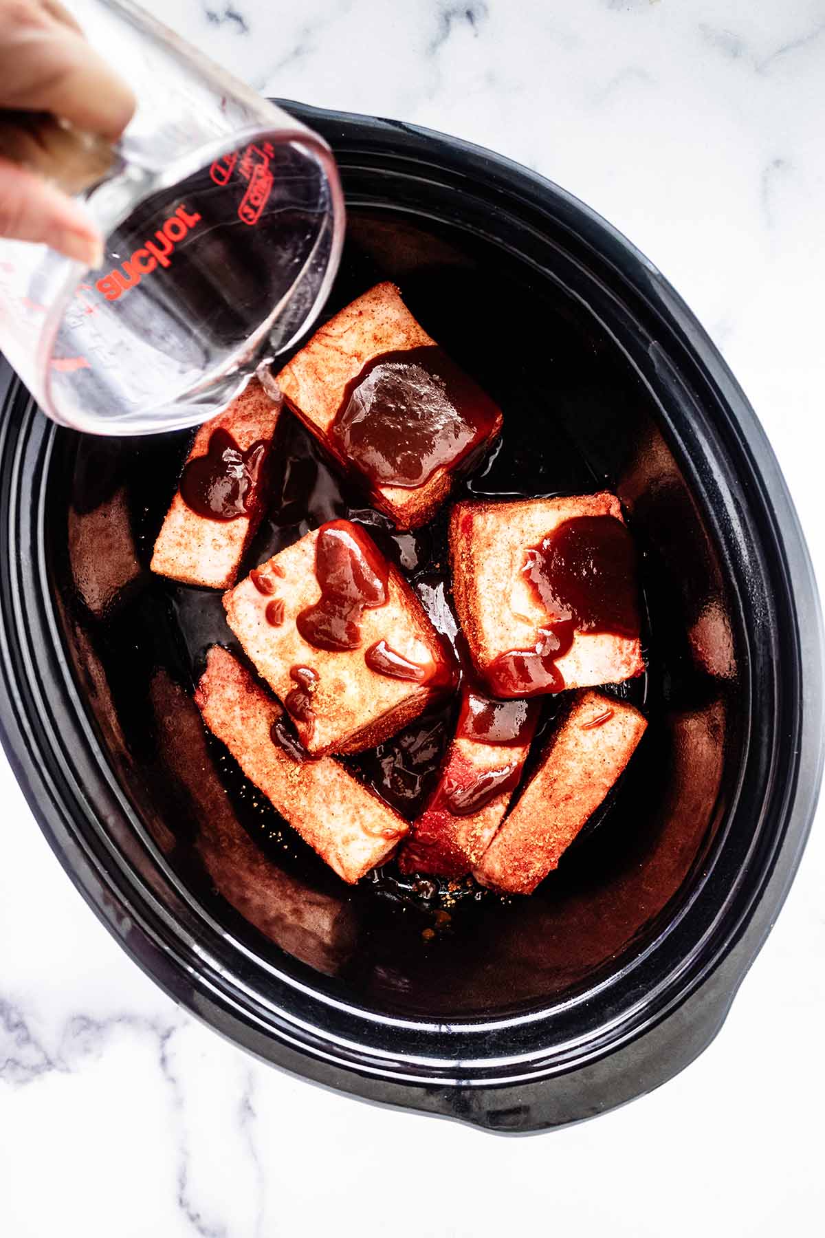 Beef short ribs in a slow cooker topped with barbecue sauce.  Water is being poured over the top.