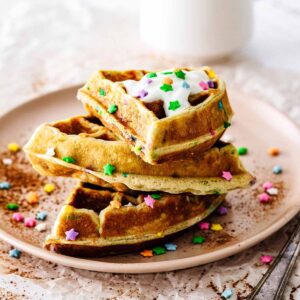Funfetti waffles stacked on a pink plate topped with whipped cream and candy sprinkles