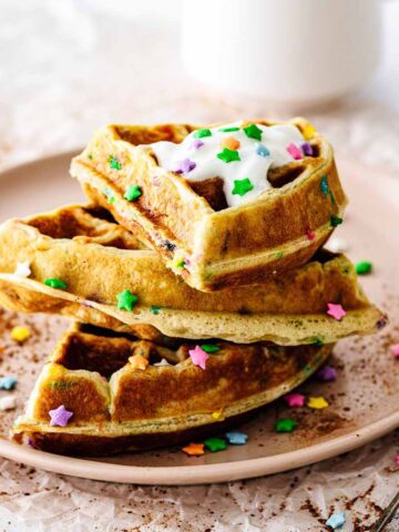 Funfetti waffles stacked on a pink plate topped with whipped cream and candy sprinkles