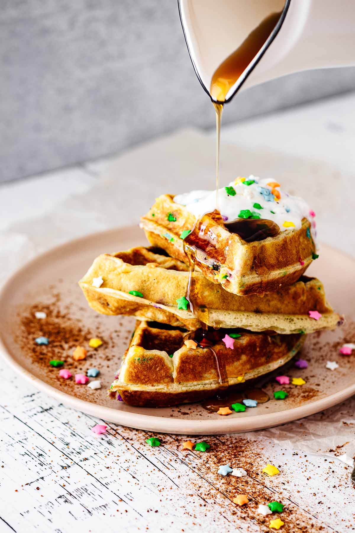 Maple syrup being poured over a stack of funfetti waffles topped with whipped cream and candy sprinkles on a pink plate.