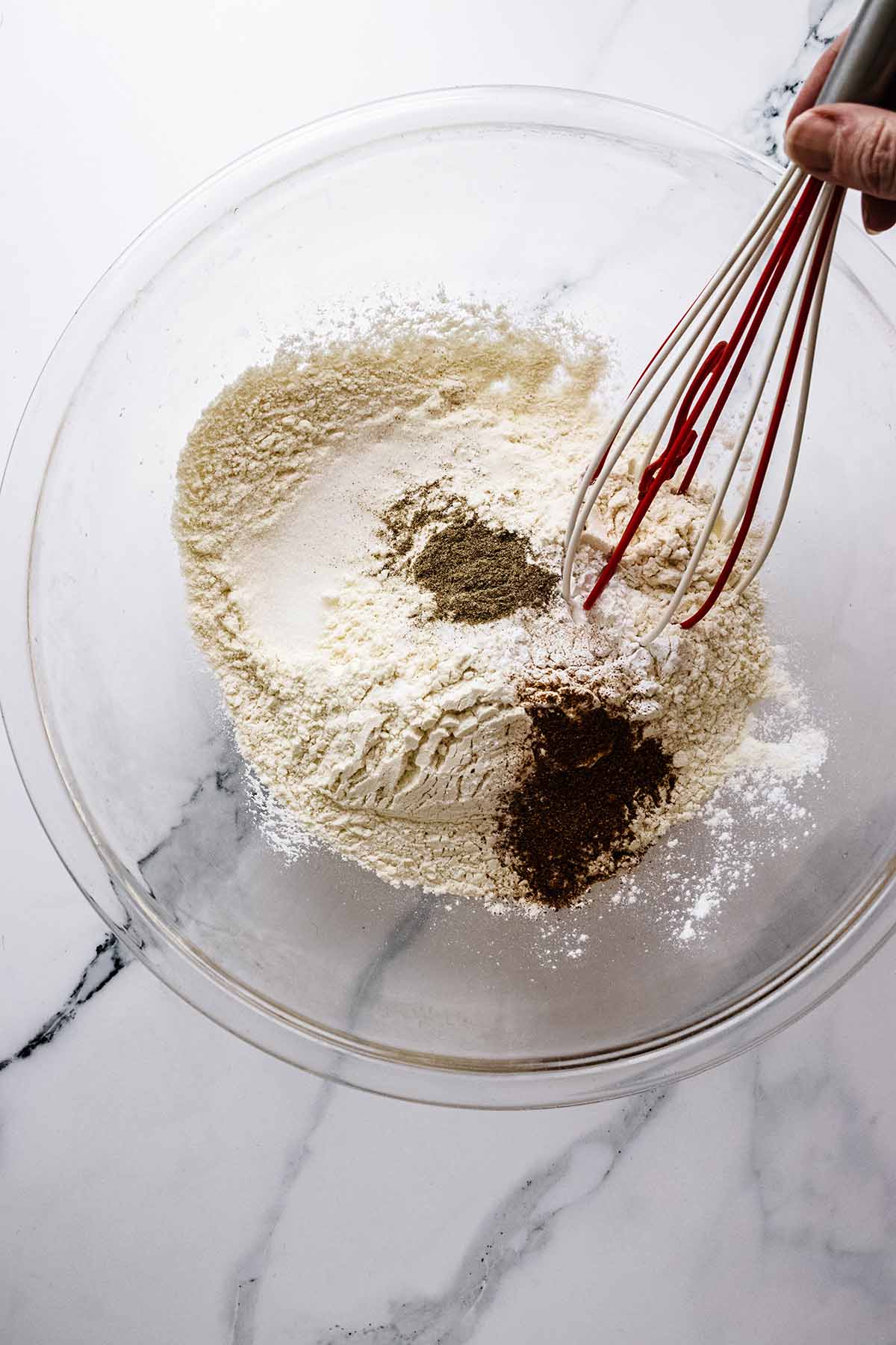 Dry ingredients in a large glass bowl on a marble countertop with a red and white whisk.