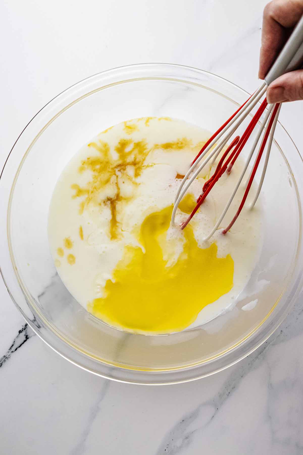 Wet ingredients in a large glass bowl on a marble countertop with a red and white whisk.