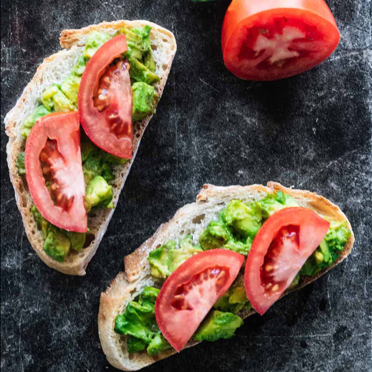 Two slices of avocado tomato toast on a dark marble background.