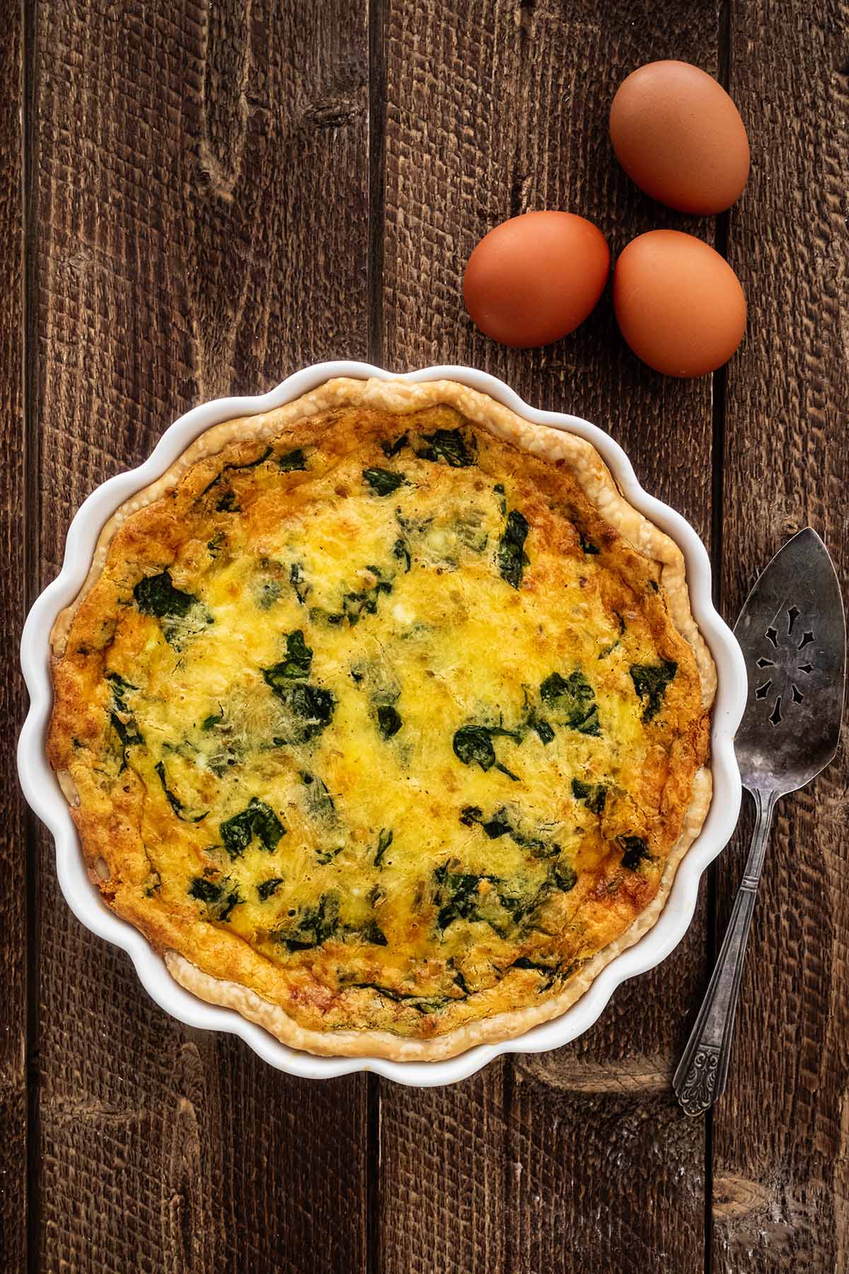 Quiche Florentine in a white quiche dish with a pie server and three eggs on a dark wood background.