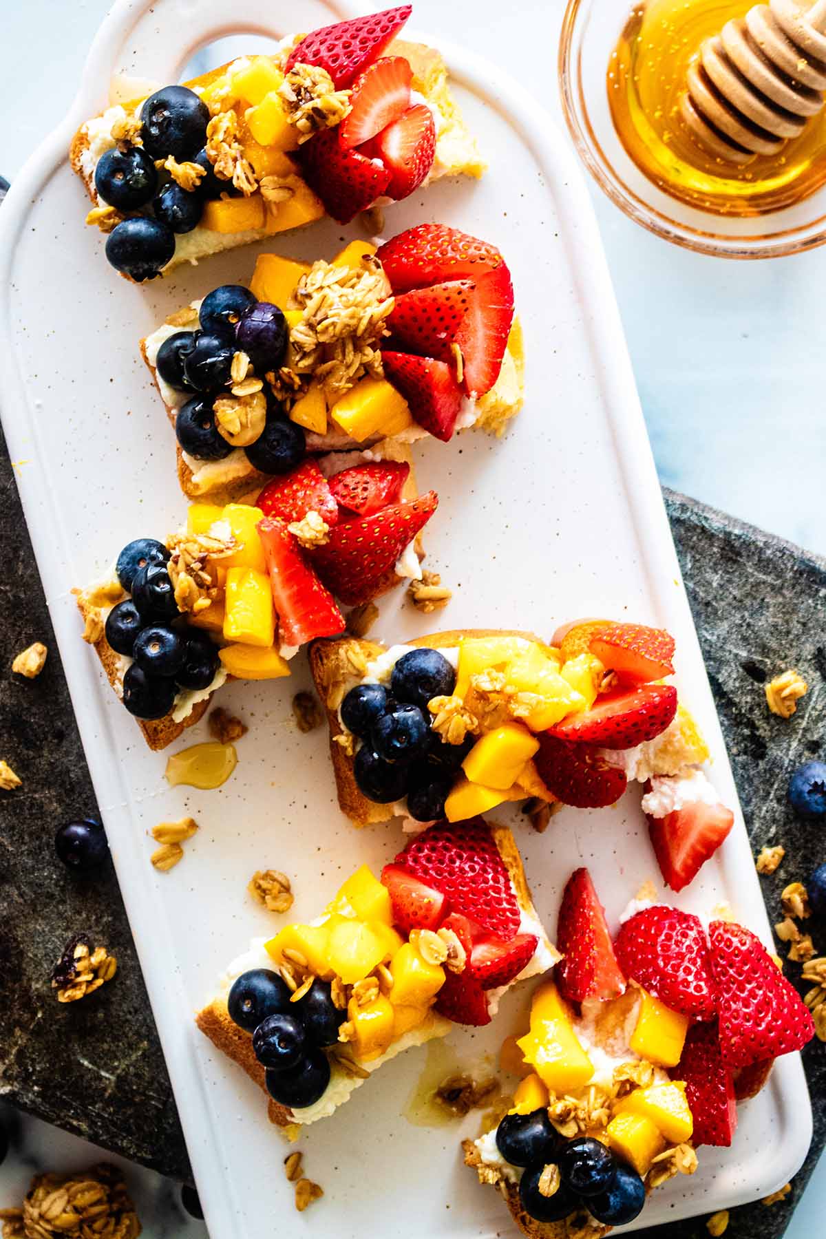 Slices of fruit-topped ricotta toast on a white ceramic serving board with a small bowl of honey and honey wand.