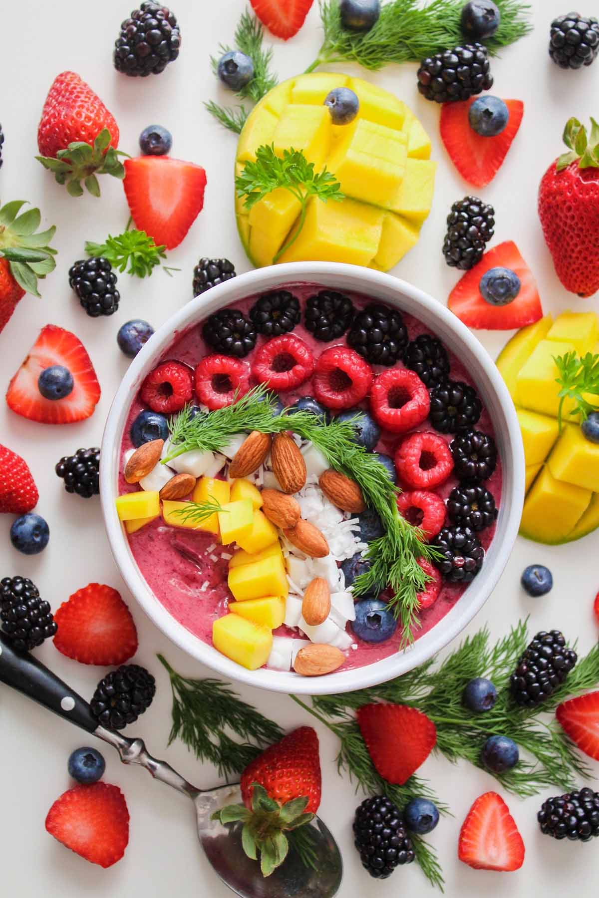 Fresh fruit on a white background surrounding a smoothie bowl topped with fresh fruit in a white bowl