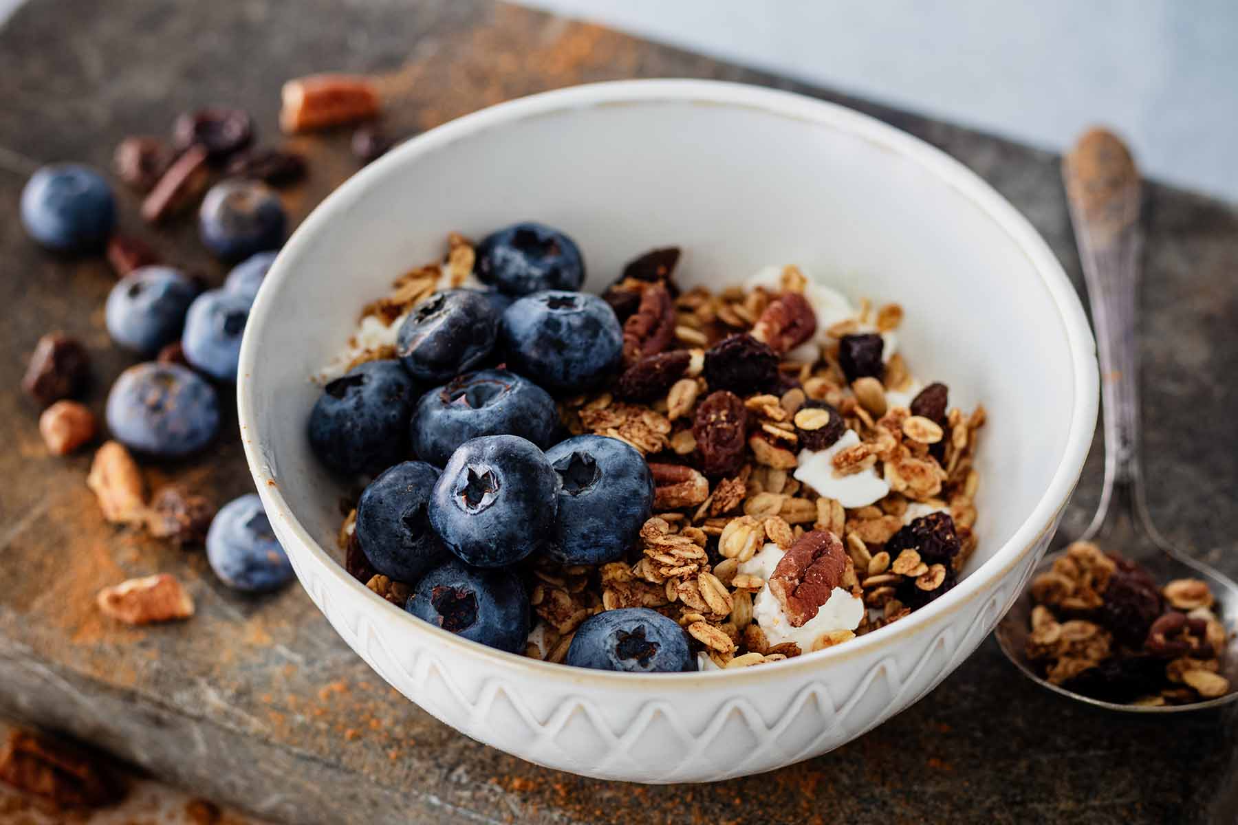 Cinnamon granola in a white ceramic bowl with fresh blueberries and a soup spoon with granola.