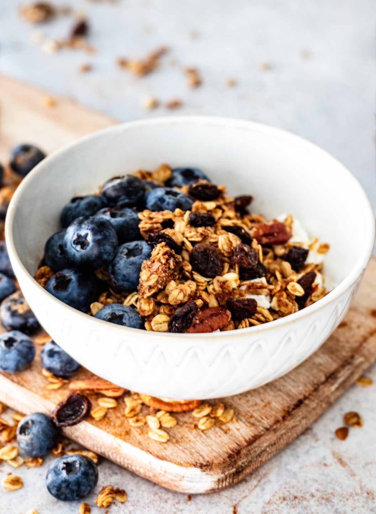 Cinnamon granola in a white ceramic bowl on a wooden cutting board with fresh blueberries
