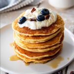 Stack of sweet cream pancakes topped with whipped cream, maple syrup, fresh blueberries, and chopped pecans on a white plate.