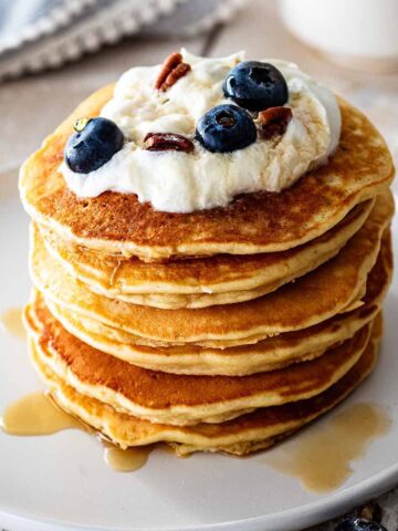 Stack of sweet cream pancakes topped with whipped cream, maple syrup, fresh blueberries, and chopped pecans on a white plate.