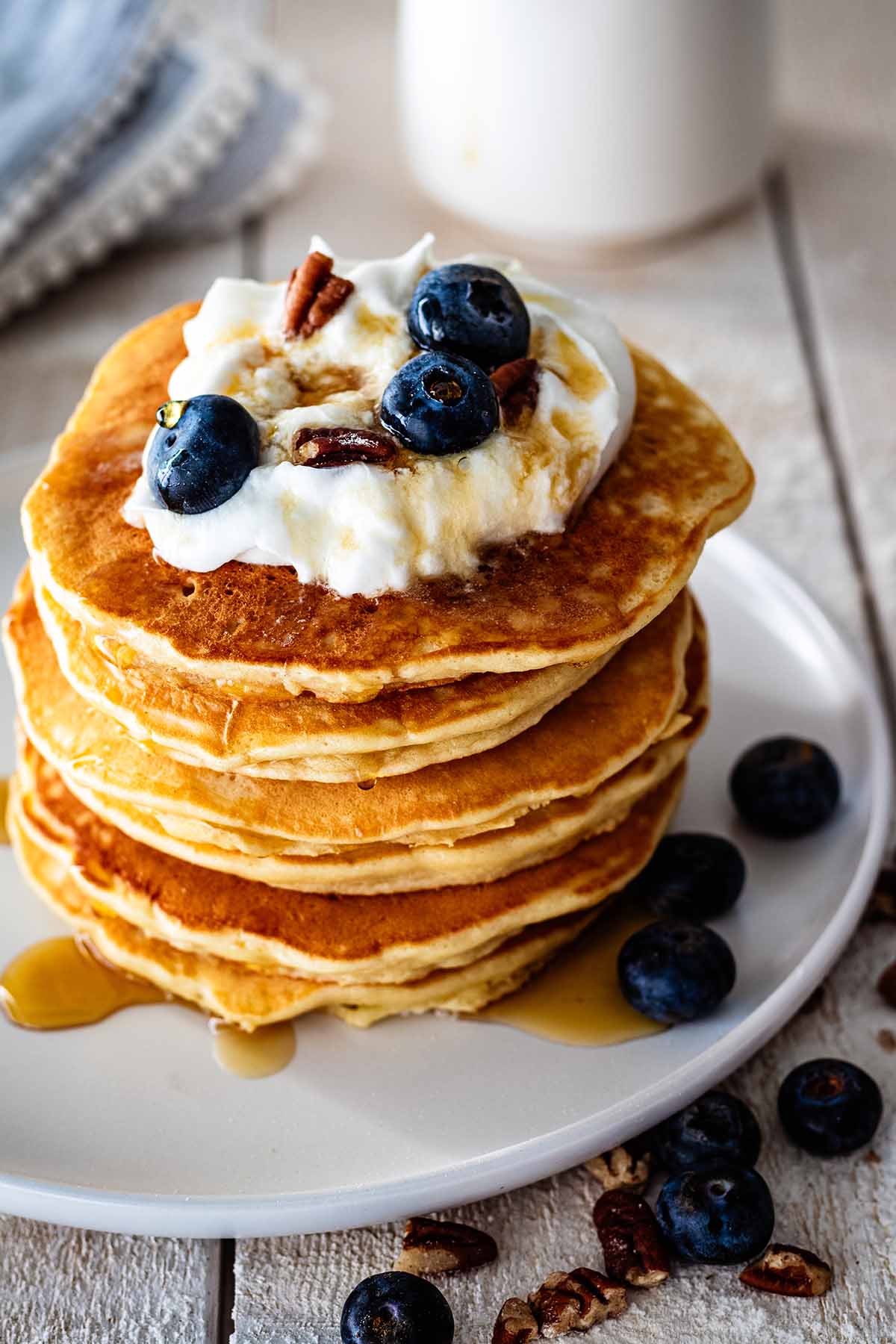 Stack of sweet cream pancakes topped with whipped cream, maple syrup, fresh blueberries, and chopped pecans on a white plate.