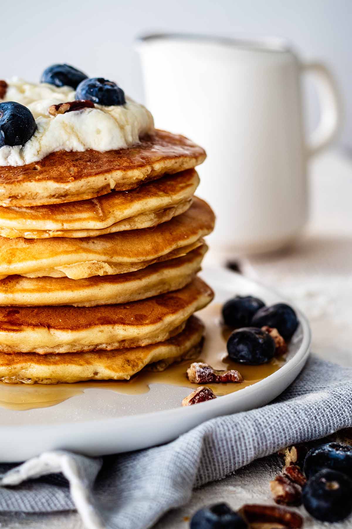 Close up of a stack of sweet cream pancakes topped with whipped cream, maple syrup, fresh blueberries, and chopped pecans on a white plate.  on top of a gray napkin.