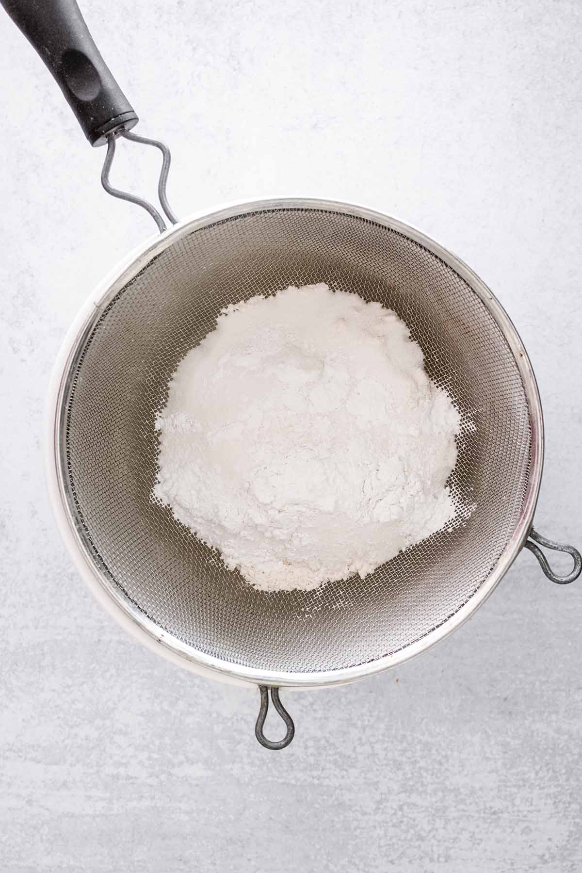 Dry ingredients in a fine mesh strainer over a white ceramic bowl