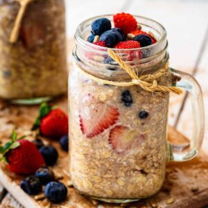 Close up of overnight oats without milk topped with sliced ​​strawberries and fresh blueberries in a glass mug jar.