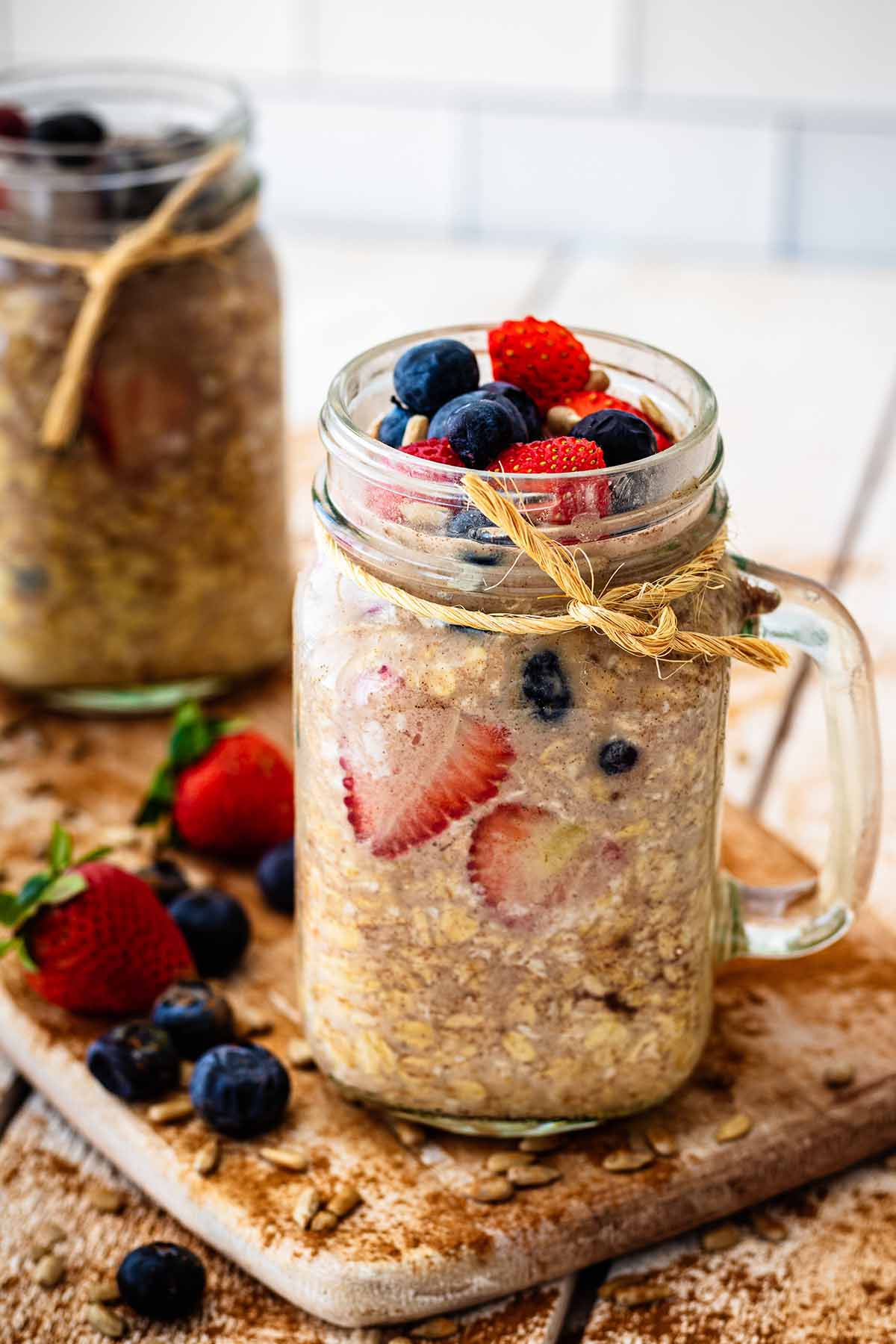 Two glass mug jars filled with overnight oats without milk topped with sliced ​​strawberries and fresh blueberries.