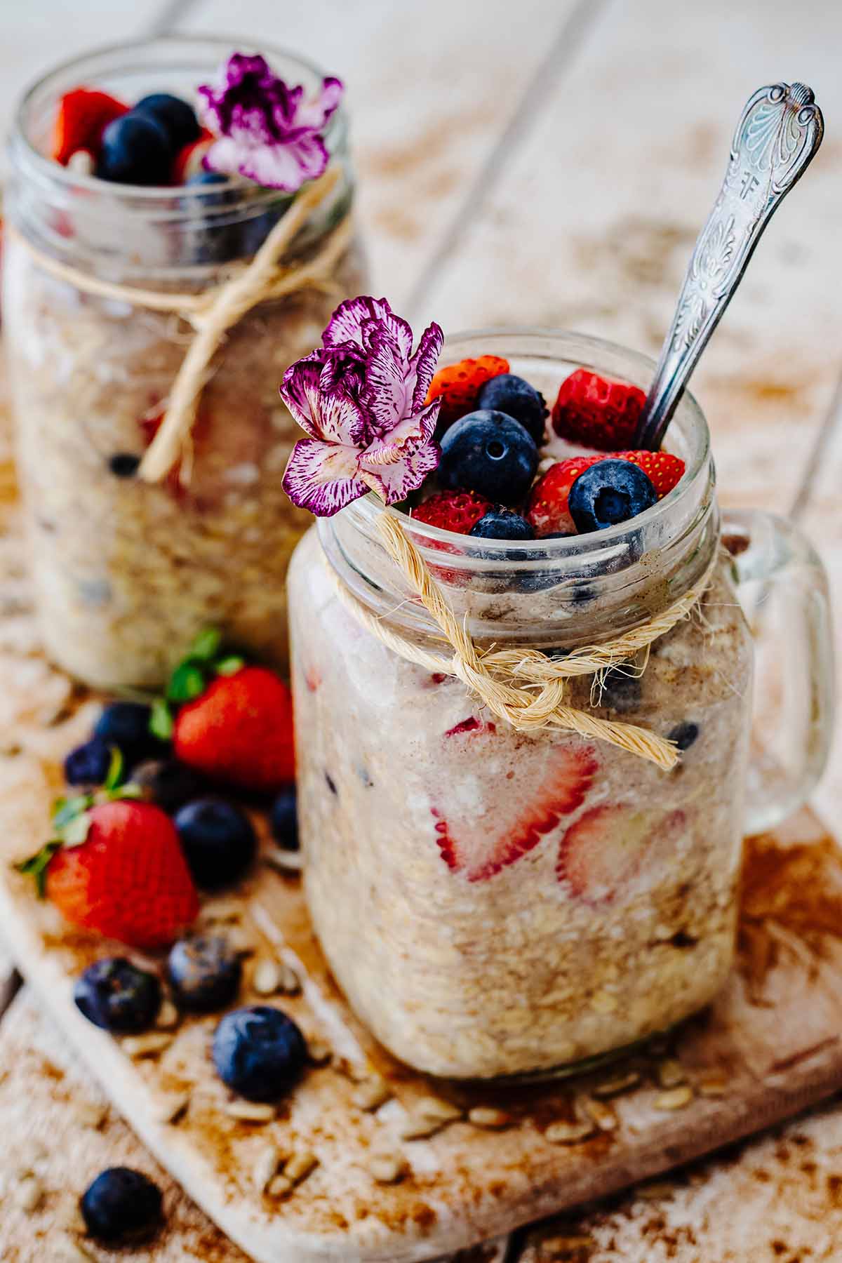 Two glass mug jars filled with overnight oats without milk topped with fresh berries and a mini purple and white carnation.