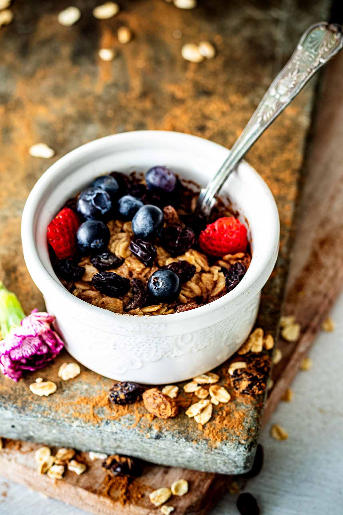 Single serving baked oatmeal topped with sliced ​​strawberries and fresh blueberries in a white ceramic ramekin with a spoon..