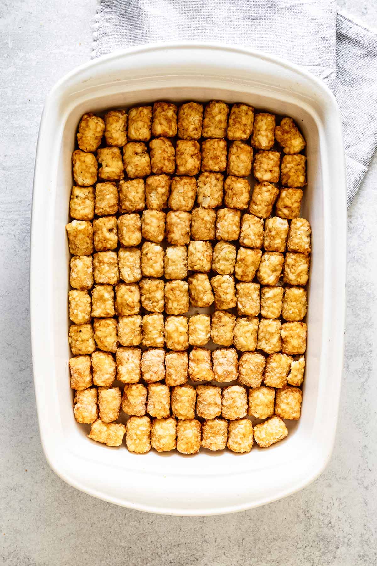 Frozen tater tots lined up at the bottom of a white ceramic baking dish.