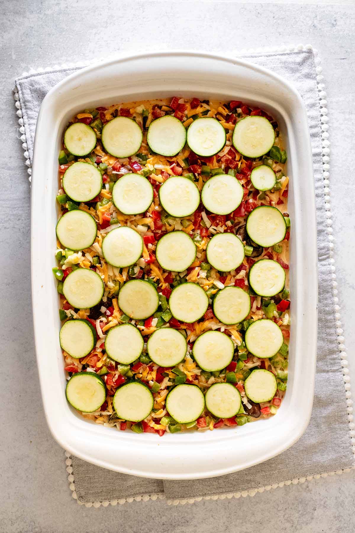 Casserole mixture topped with zucchini slices in a white ceramic baking dish.