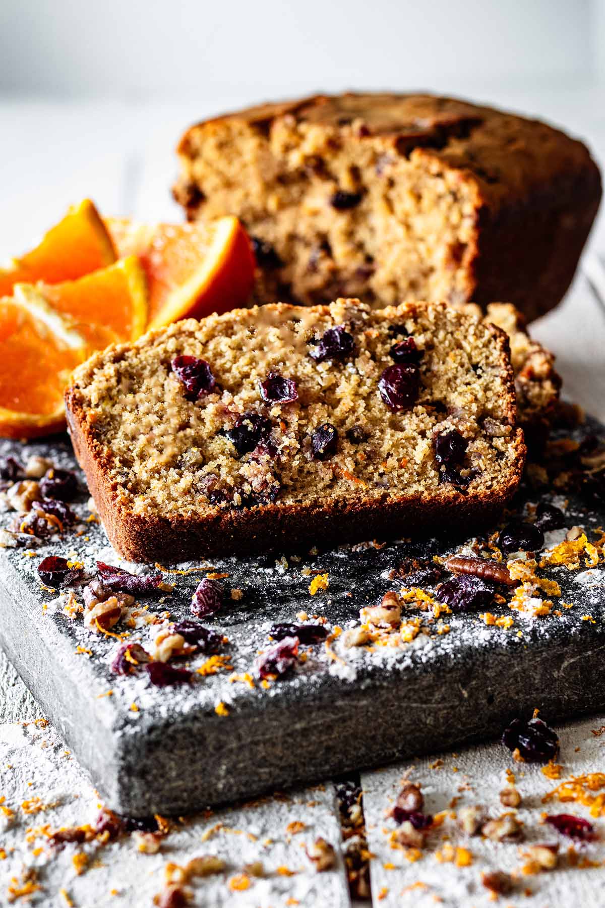 Close up of a slice of cranberry orange bread with sliced ​​oranges and a loaf of sweet bread.
