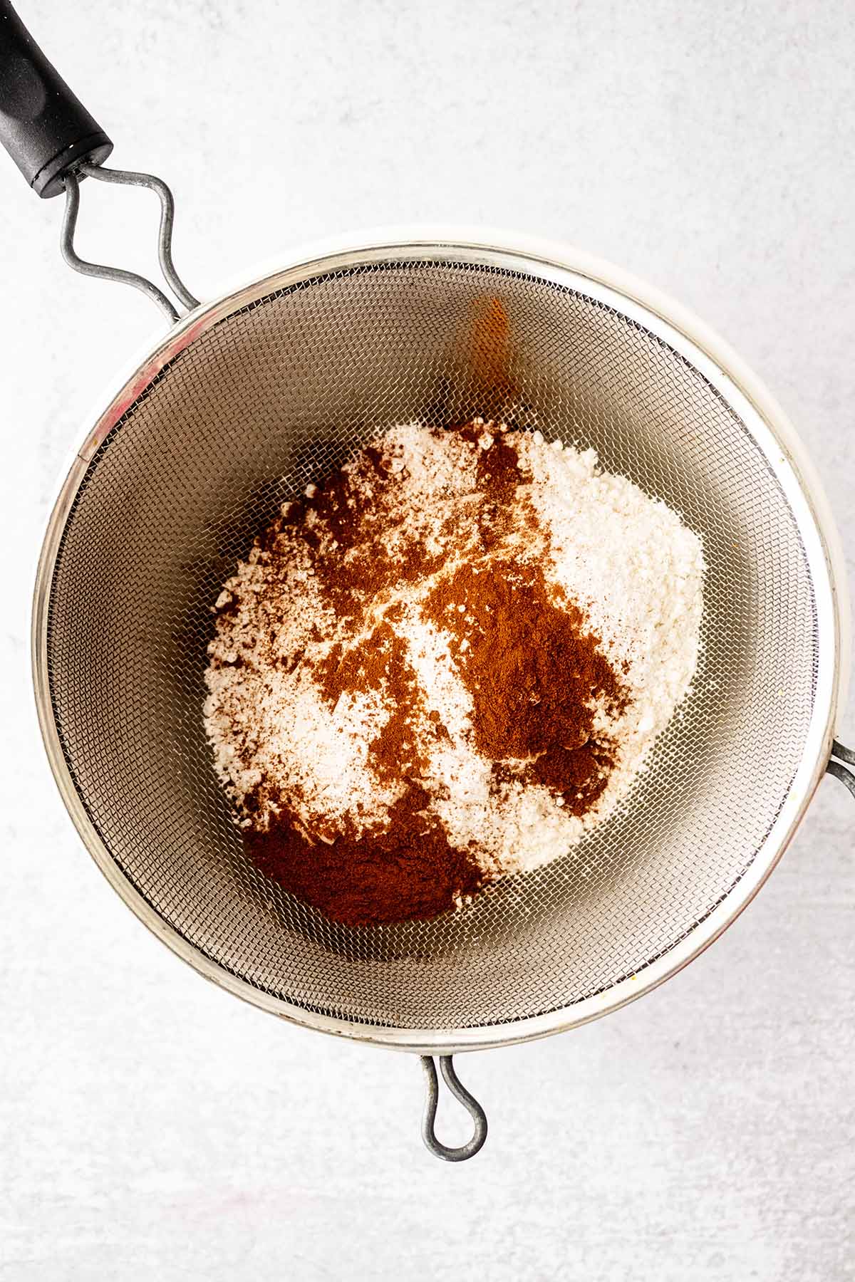 Dry ingredients in a mesh strainer on top of a white ceramic bowl.
