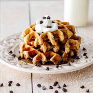 Stack of chocolate chip waffles topped with whipped cream, chocolate chips, and maple syrup on a white plate.
