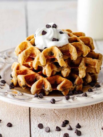 Stack of chocolate chip waffles topped with whipped cream, chocolate chips, and maple syrup on a white plate.
