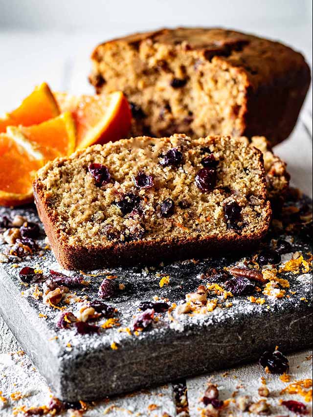 Slice of cranberry orange bread on a cutting board with orange slices and sliced loaf