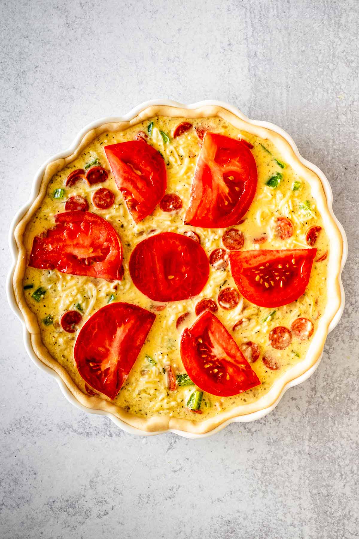 Unbaked Italian quiche topped with sliced ​​tomatoes in a white quiche dish.