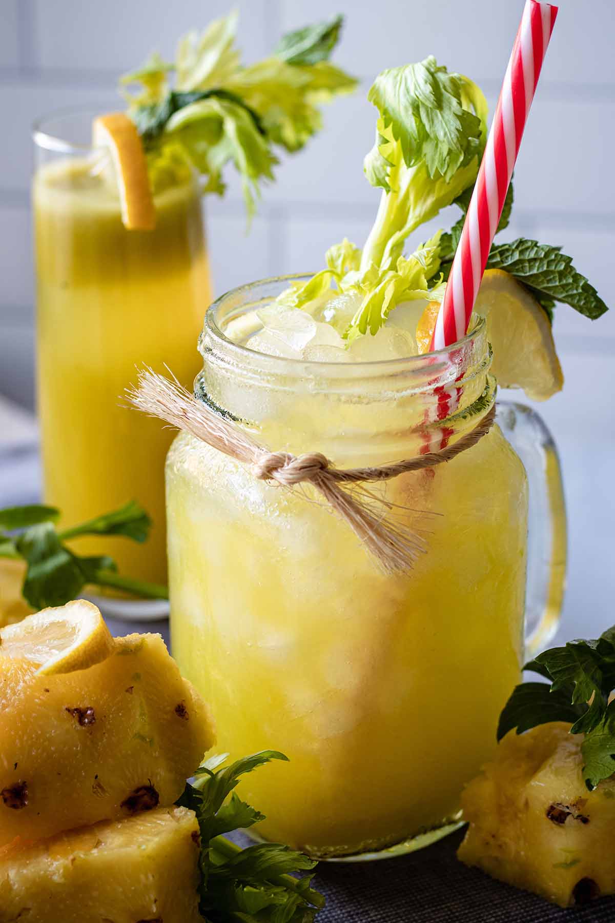Close up of pineapple celery juice with a red and white straw garnished with celery greens, fresh mint, and a lemon slice in a mug jar .