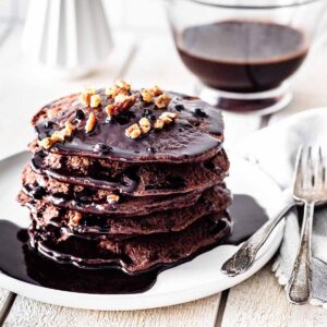 Stack of chocolate protein pancakes topped with chocolate pancake syrup and chopped pecans on a white plate.