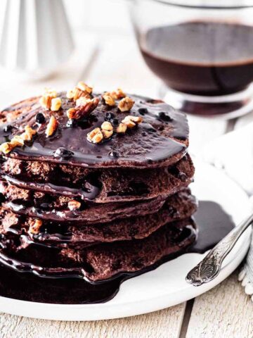 Stack of chocolate protein pancakes topped with chocolate pancake syrup and chopped pecans on a white plate.