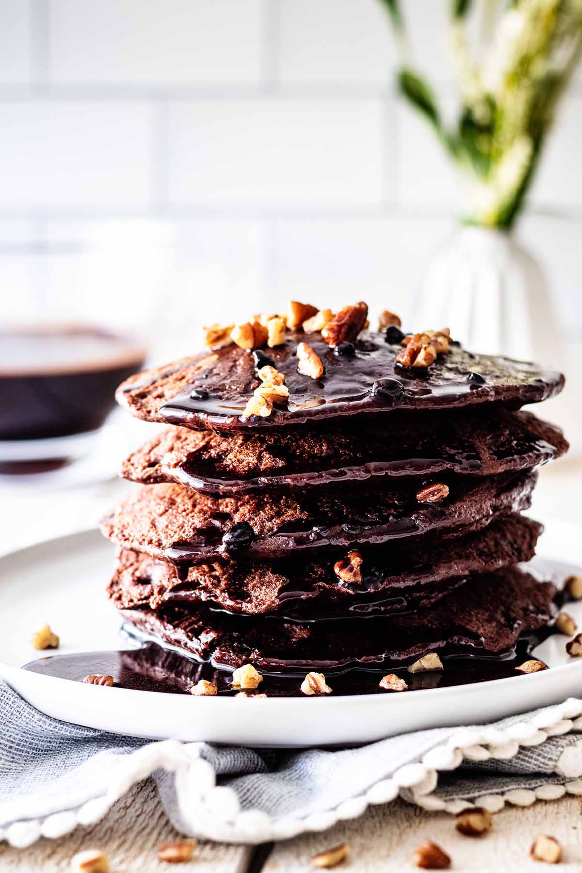 Stack of chocolate protein pancakes topped with chocolate pancake syrup and chopped pecans on a white plate.