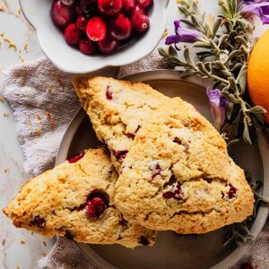 Three orange cranberry scones on a light grey plate with flowers and a bowl of fresh cranberries.