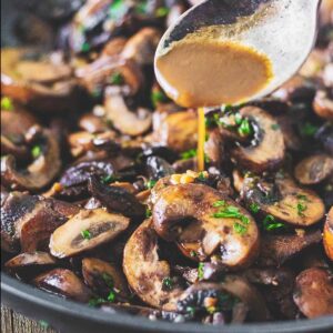 Sauce from the skillet being spooned over sautéed garlic butter mushrooms as they sit in the skillet.
