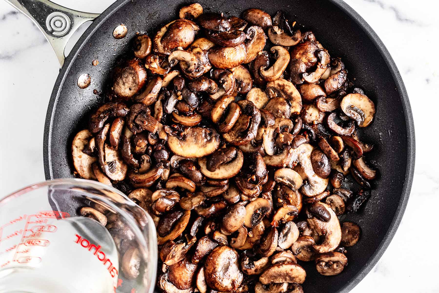 White wine being poured over sautéed sliced mushrooms in a skillet.