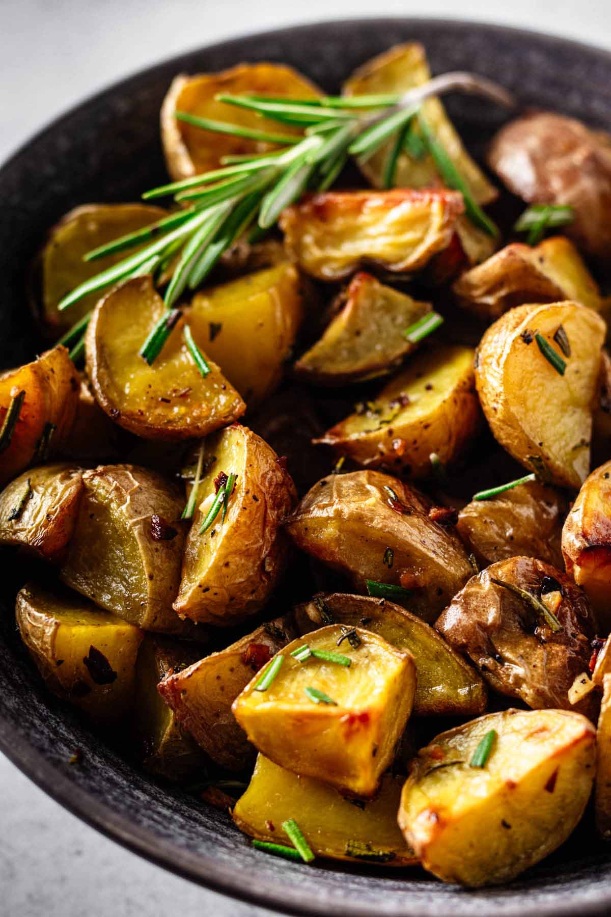 Close up of garlic rosemary roasted potatoes in a dark grey bowl with a sprig of rosemary.