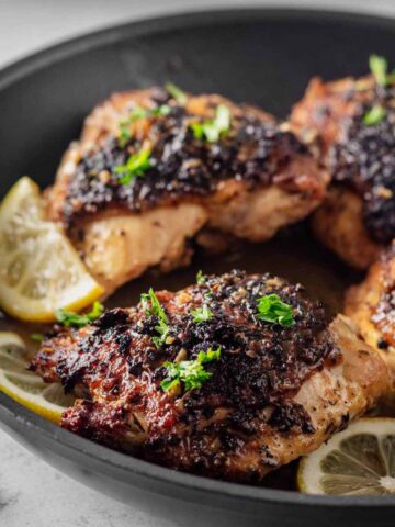 Close up of four cooked lemon pepper chicken thighs garnished with chopped parsley in a skillet with lemon slices.