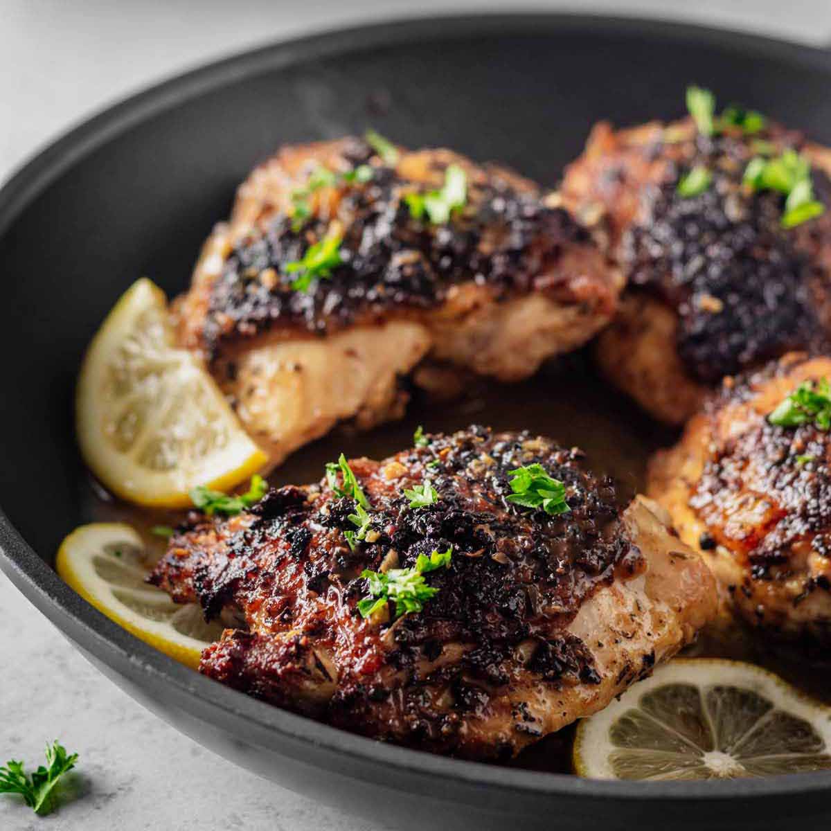 Close up of four cooked lemon pepper chicken thighs garnished with chopped parsley in a skillet with lemon slices.