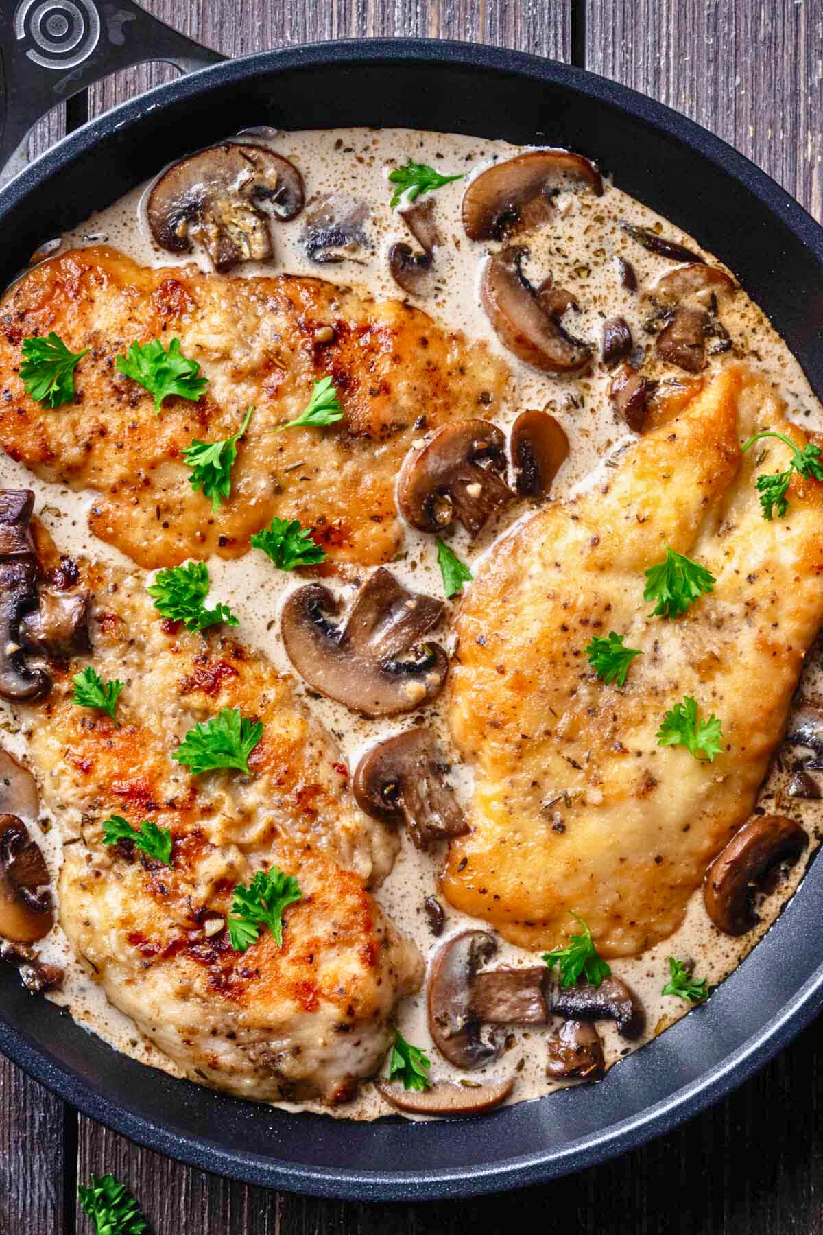 Closeup overhead view of three cooked chicken cutlets in mushroom sauce topped with chopped parsley in a large skillet